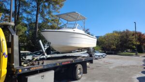 A white boat is loaded on a truck bed, surrounded by trees and parked cars under a clear blue sky.