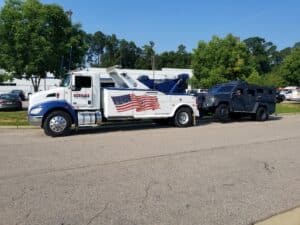 Tow truck with American flag design towing a black military-style vehicle on a sunny day.