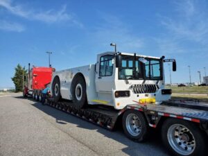 A white utility vehicle is loaded on a flatbed truck, with a red truck in the background.