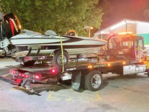 Tow truck transporting a Triton bass boat at night, illuminated by truck lights.
