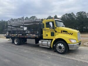 Yellow towing truck with a ladder rack parked on a gravel road surrounded by trees.