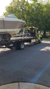 A flatbed tow truck carries a boat, parked on a tree-lined street in daylight.