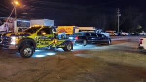 Tow truck with bright colors is parked next to a black limousine at night in a crowded lot.