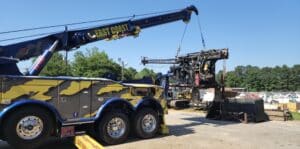 A blue and yellow tow truck lifts heavy equipment at a construction site with clear skies.